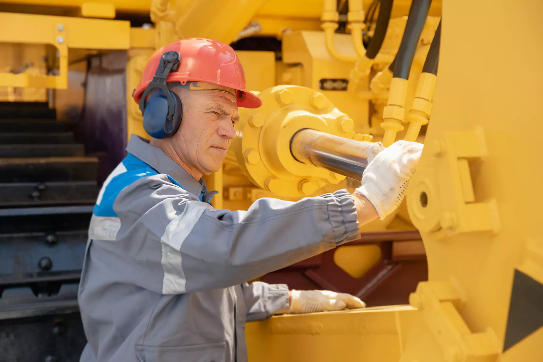 A worker wearing a red hard hat, ear protection, and gloves operates machinery, focusing on a hydraulic component.